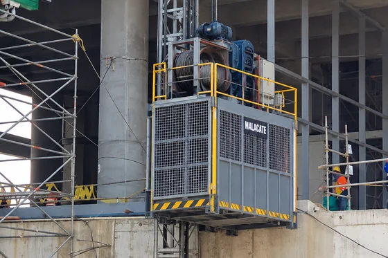 Malacate de obra con cabina de carga, motor reductor y tambor con cable sobre un mástil en una edificación en construcción